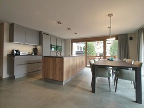 Eine moderne Küche mit einer zentralen Kochinsel, einem Essbereich mit Tisch und Stühlen sowie einem Ausblick durch große Fenster.