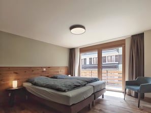 A bedroom with a wooden headboard, a bed with grey bedding, a bedside lamp, a chair, and a view of a balcony with wooden railings. - Derby Appartement/Fewo 8-Bettwohnung by Interhome (Saas-Fee)