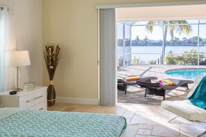 A bedroom with a bed, a nightstand, a lamp, and a vase with dried plants. There is a sliding door leading to a balcony with a pool, lounge chairs, and a view of a body of water. - Villa in Cape Coral with swimming pool (Cape Coral)