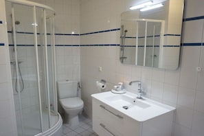 Casa de banho com chuveiro em cabine de vidro, sanita, lavatório com espelho e paredes revestidas a mosaico branco.