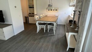 Un comedor con una mesa de madera y sillas blancas, una lámpara colgante y un armario blanco con estantes.