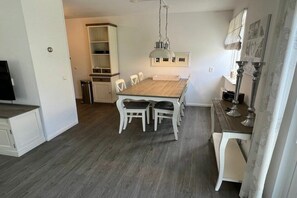 Uma área de refeições com mesa de madeira e cadeiras brancas, luminária pendente e armário branco com prateleiras.