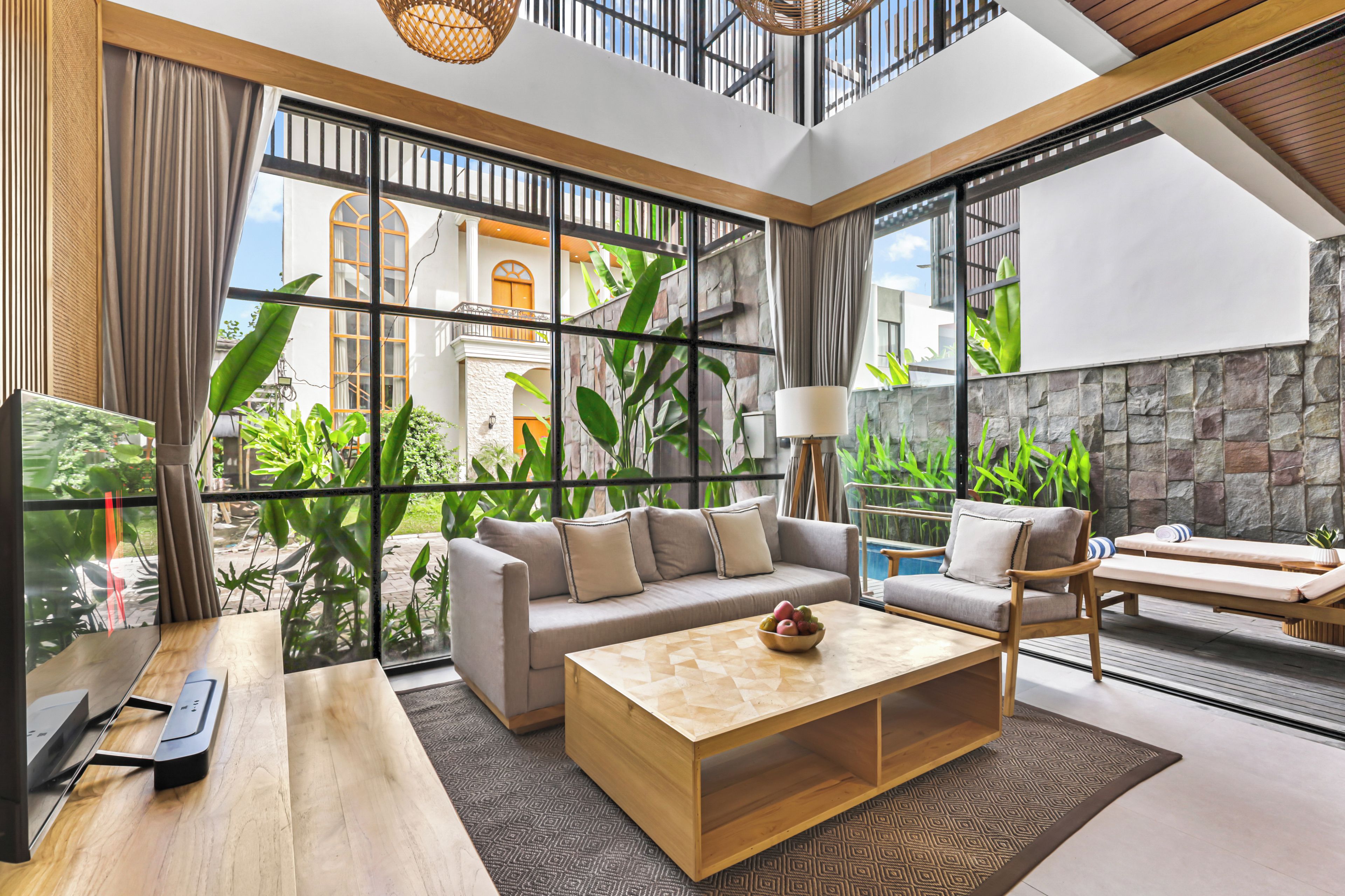Una sala de estar moderna con sofá, mesa de centro y sillones, ventanas amplias con vistas a un patio y un edificio, además de una pared de piedra.