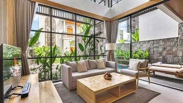 Una sala de estar moderna con sofá, mesa de centro y sillones, ventanas amplias con vistas a un patio y un edificio, además de una pared de piedra.