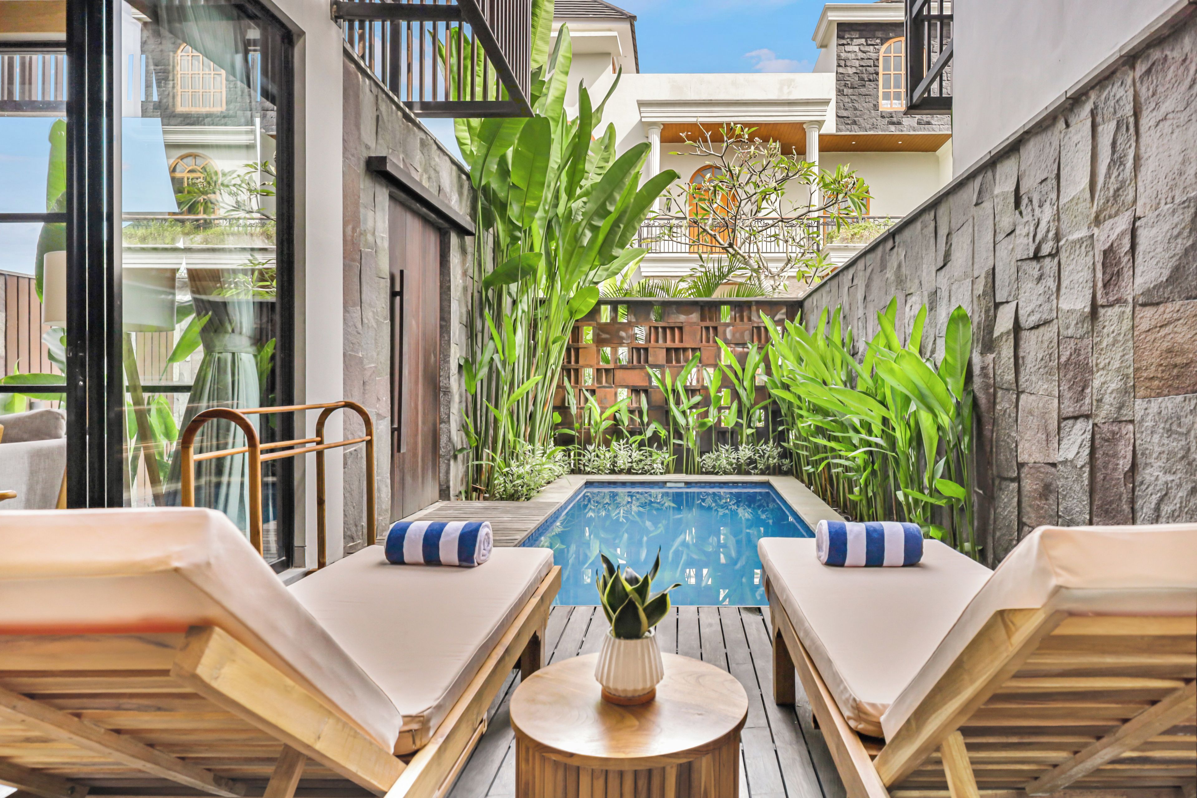 Un espace aménagé au bord de la piscine, avec des chaises longues et une petite table, entouré de verdure et de murs en pierre.