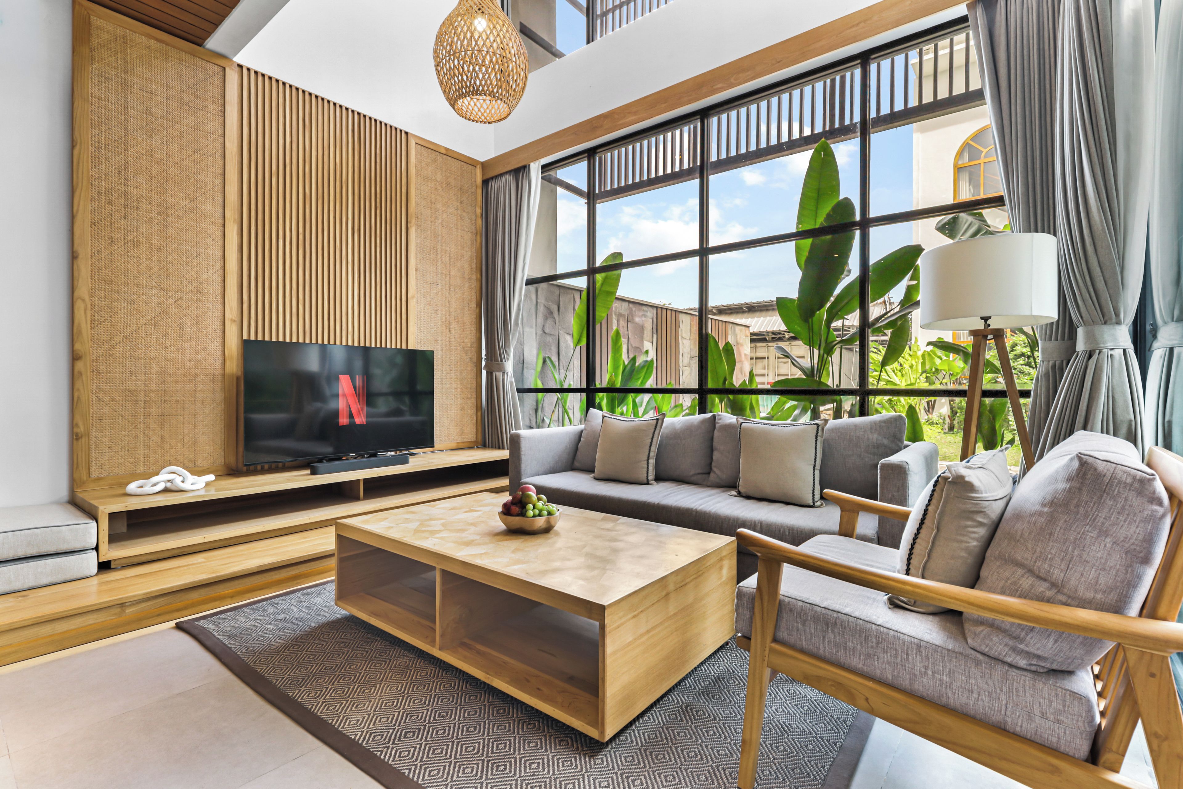 A modern living room with a television, sofa, coffee table, and large windows with plants.