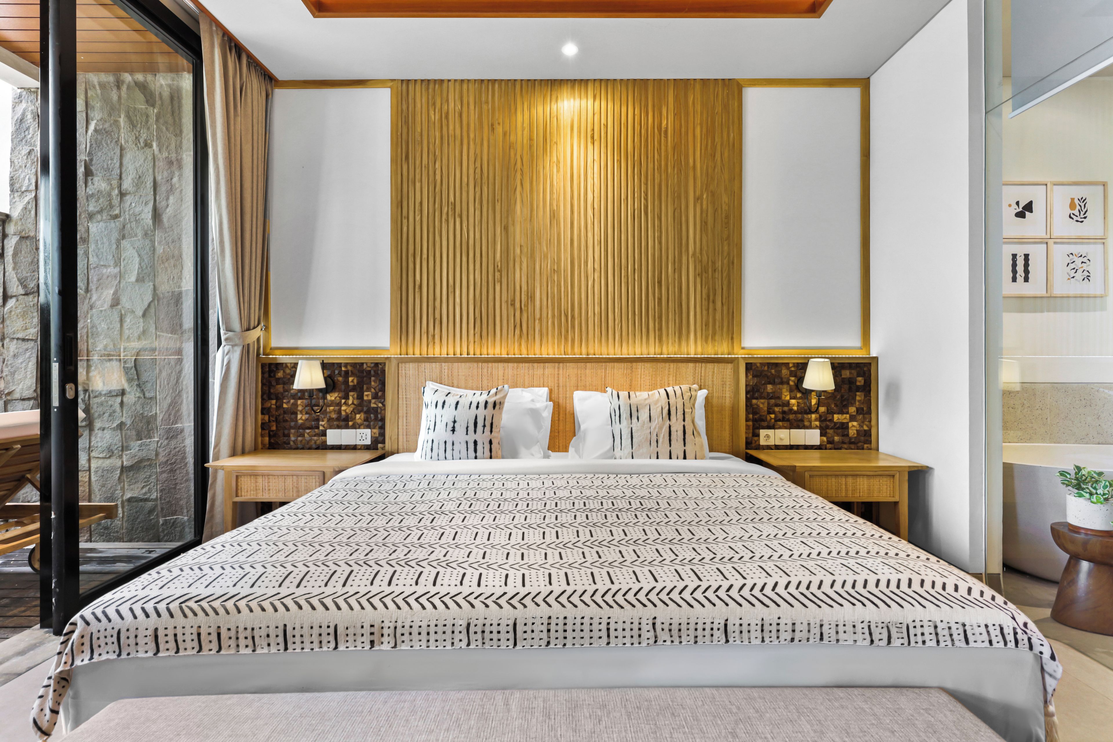 Een moderne hotelkamer met een groot bed, twee nachtkastjes met lampen, een houten hoofdbord en uitzicht door een glazen deur.