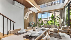 Un espace repas moderne avec une longue table en bois dressée pour un repas, entourée de sièges confortables et de grandes fenêtres offrant une vue sur la verdure.