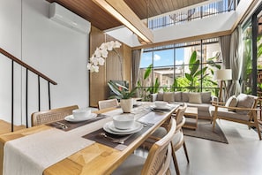 A modern dining area with a long wooden table set for a meal, surrounded by comfortable seating and large windows offering a view of greenery. - Cardano Villa by Kozystay - Canggu (North Kuta, Badung)