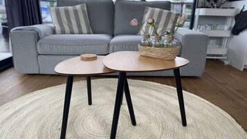 Ein modernes Wohnzimmer mit einem grauen Sofa, einem runden Couchtisch mit schwarzen Beinen und einem gewebten Korb mit Blumen.