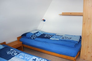 Ein Einzelbett mit blauer Matratze und gemusterten Betttextilien, ein Nachttisch aus Holz und ein an der Wand befestigtes Regal.