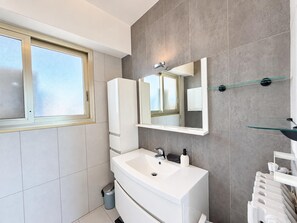 Ein modernes Badezimmer mit großem Fenster, Waschbecken in Weiß, einem Spiegel und Ablagefläche aus Glas.