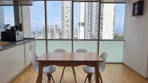 Een eethoek met een houten tafel en witte stoelen, grote ramen met uitzicht over de stad, en een keuken met een koffieapparaat.