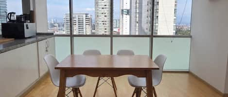 Un coin repas avec une table en bois et des chaises blanches, de grandes fenêtres offrant une vue sur la ville, et un espace cuisine équipé d’une machine à café.