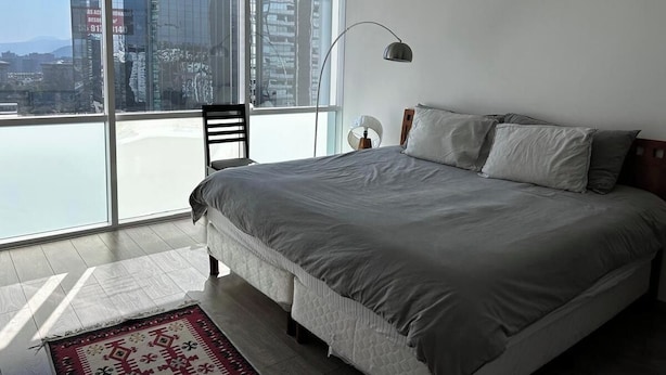 Een slaapkamer met een groot bed, een stoel, een nachtkastje en uitzicht op een stadsgezicht door middel van kamerhoge ramen.
