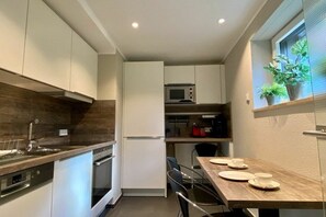 A modern kitchen with white cabinets, a built-in oven, and a microwave. There is a dining table with plates and cups, and a window with plants on the sill. - Terrace apartment garden oasis (Lindau)