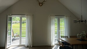Une pièce avec un plafond en pente, un crâne de cerf accroché au mur, une table à manger avec des chaises, et une vue sur un espace extérieur accessible par de grandes portes-fenêtres coulissantes, comprenant une pelouse et un fauteuil.