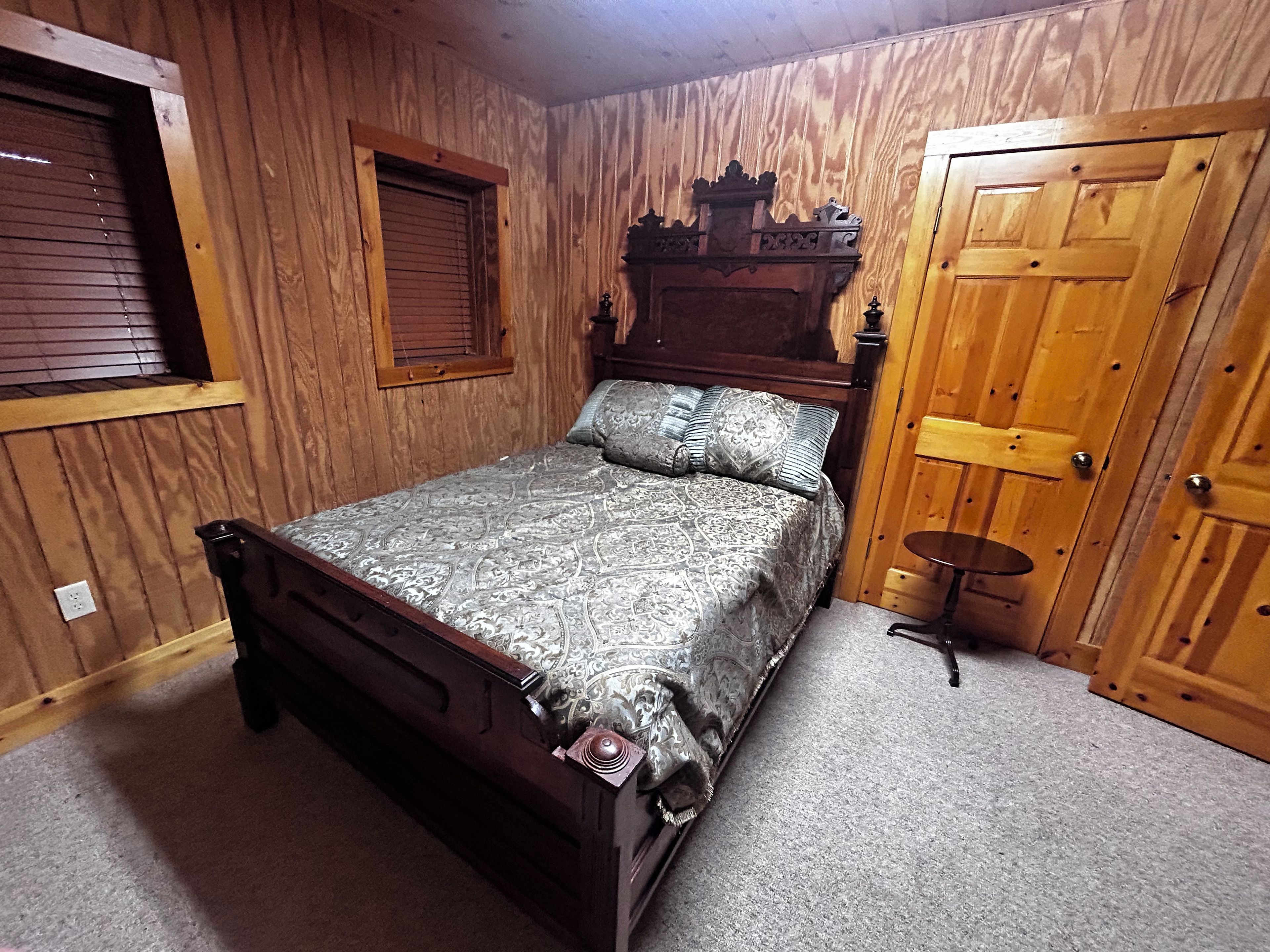 A bedroom with a wooden bed, a window with blinds, a wooden door, and a small round table.