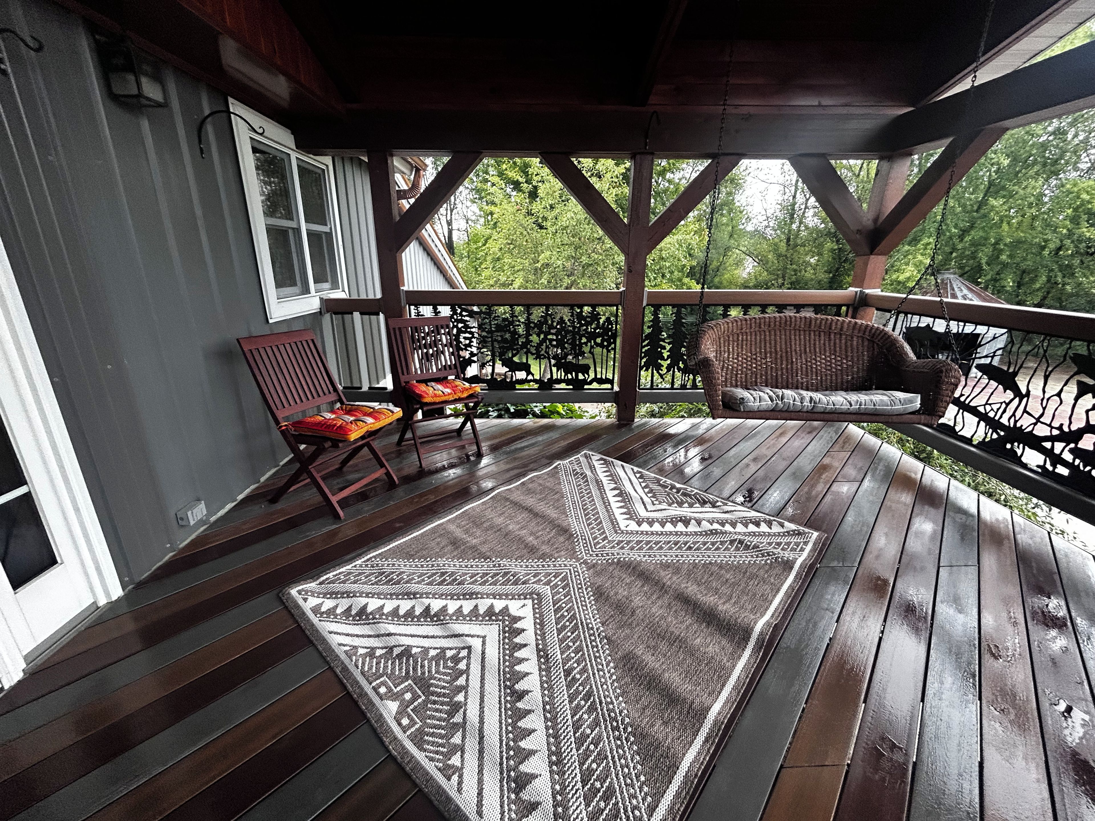 A covered porch with a wooden floor, a patterned rug, two wooden chairs, and a wicker sofa.