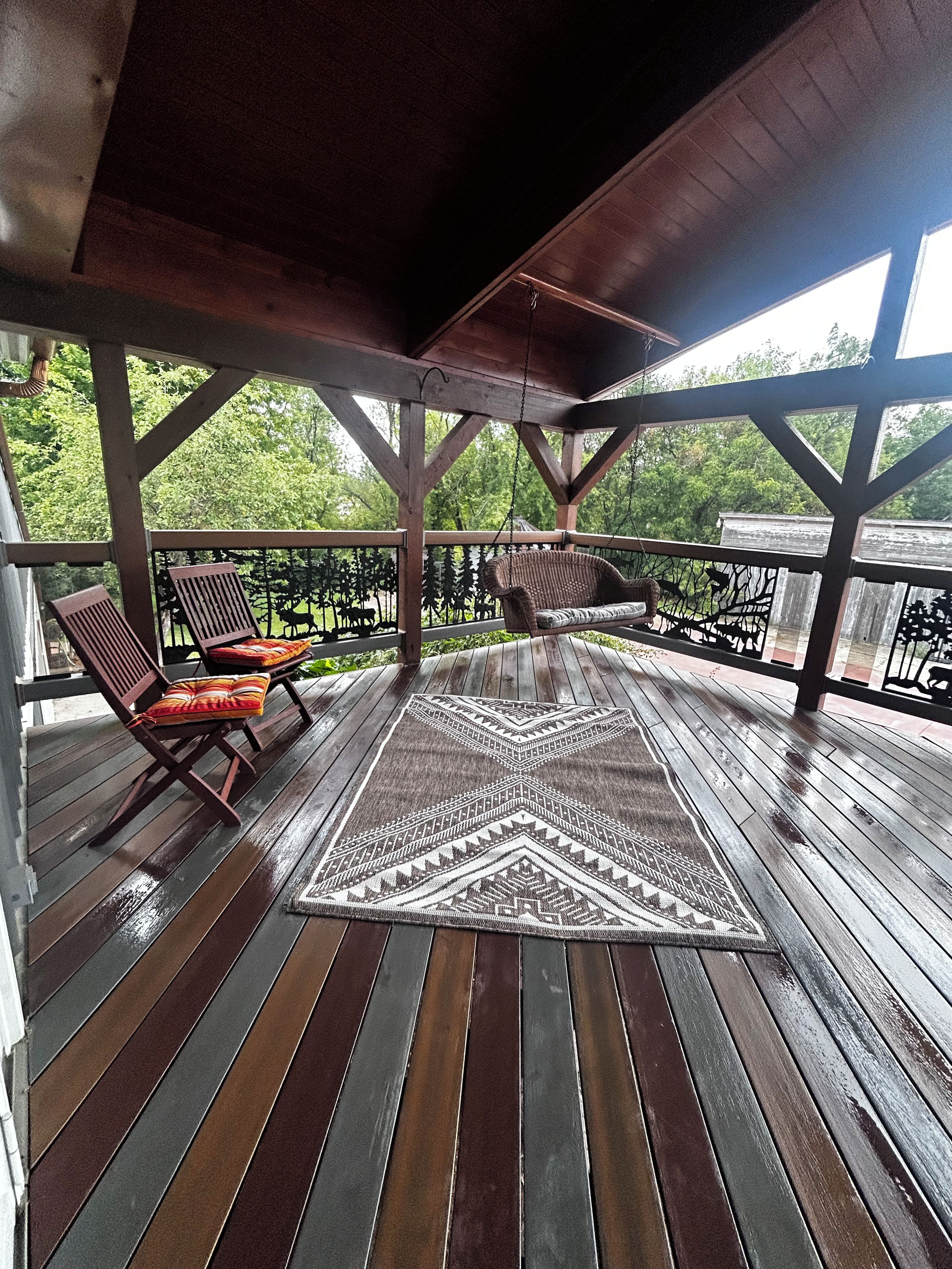 A covered wooden deck with a patterned rug, two wicker chairs, and a view of greenery.