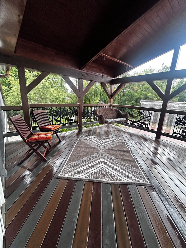A covered wooden deck with a patterned rug, two wicker chairs, and a view of greenery.