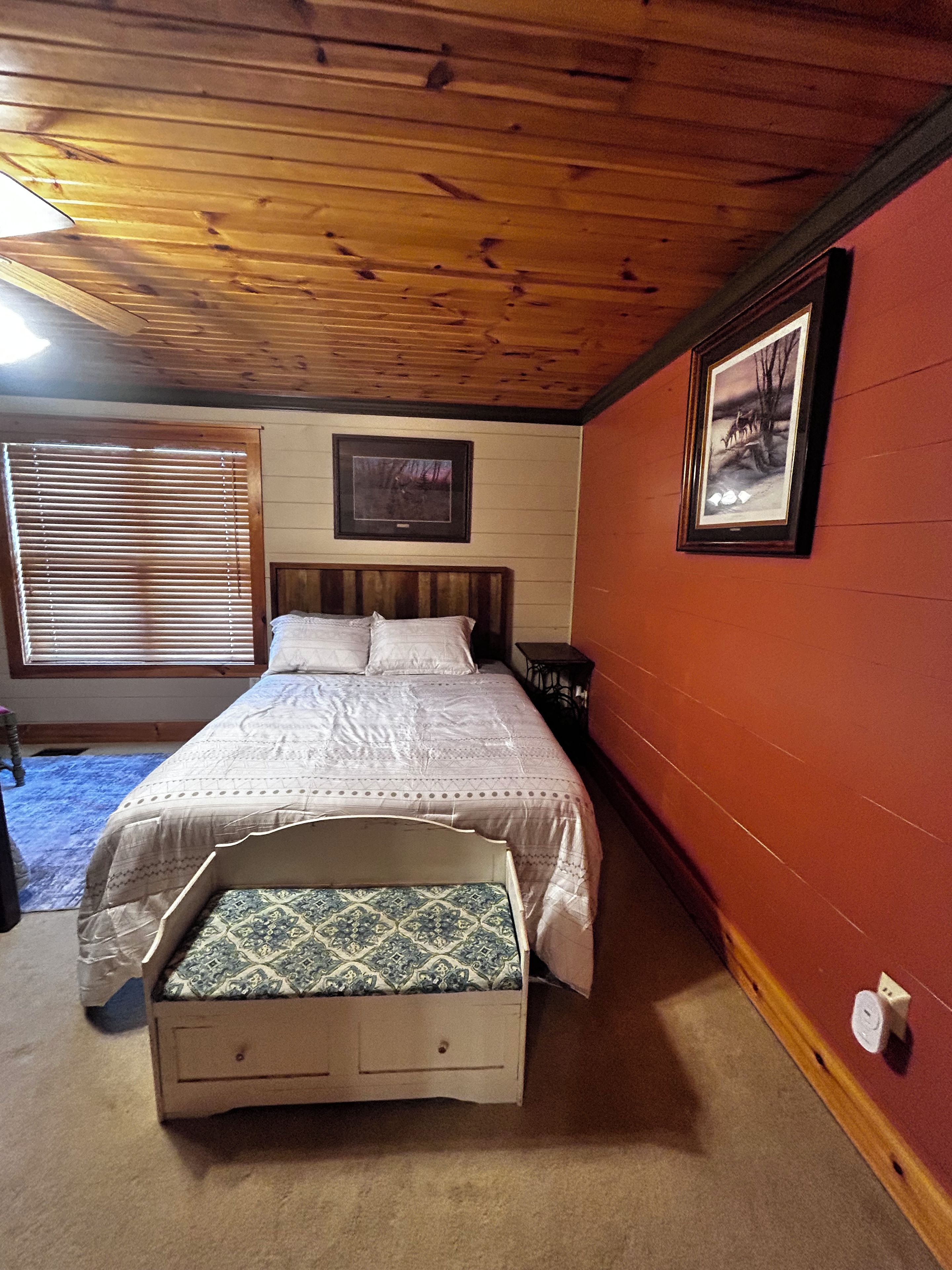 A bedroom with a bed, a bedside table, a window with blinds, a framed picture on the wall, and a red wall.