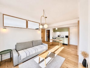 Un salon moderne avec un canapé, une table basse et un coin repas comprenant une table et des chaises. On aperçoit au fond une cuisine équipée de placards et d’appareils électroménagers.