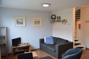 A living room with a grey sofa, a TV on a wooden stand, a small table, and a staircase leading to an upper floor. - Holiday apartment with 1 bedroom (Nes Ameland)