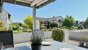 Un patio al aire libre con una mesa preparada para dos personas, rodeado de follaje y edificios residenciales al fondo.