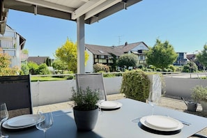 Une terrasse extérieure avec une table dressée pour deux personnes, entourée de verdure et avec des immeubles d’habitation en arrière-plan.
