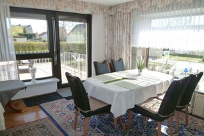 Ein Essbereich mit Tisch für zwei Personen, Stühlen, einem Fenster mit Vorhängen und Blick ins Freie.
