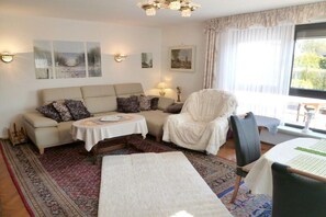 En stue med sofa, lænestol og et lille bord med lampe. Væggene er prydet med billeder, og et stort vindue er dækket af gardiner.