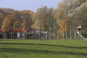 Ein Spielplatz mit Klettergerüsten und einer Rutsche, umgeben von Bäumen und einem Gebäude mit rotem Dach.