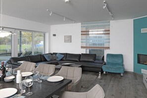 Ein modernes Wohnzimmer mit Sofa, Esstisch und Stühlen, Kamin und großem Fenster mit Jalousien.