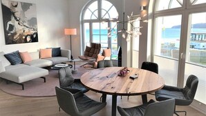 Moderni olohuone, jossa on kulmasohva, pyöreä puupöytä ja suuret ikkunat, joista avautuu näkymä merelle.
