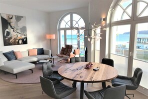 Un soggiorno moderno con un divano componibile, un tavolino rotondo in legno e vista mare attraverso ampie finestre.