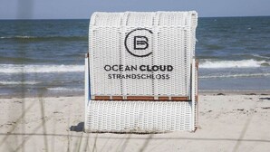 Rannalle levitetty pyyhe, jossa on painatus «OCEAN CLOUD STRANDSCHLOSS», ja taustalla näkyy hiekkaranta ja meri.