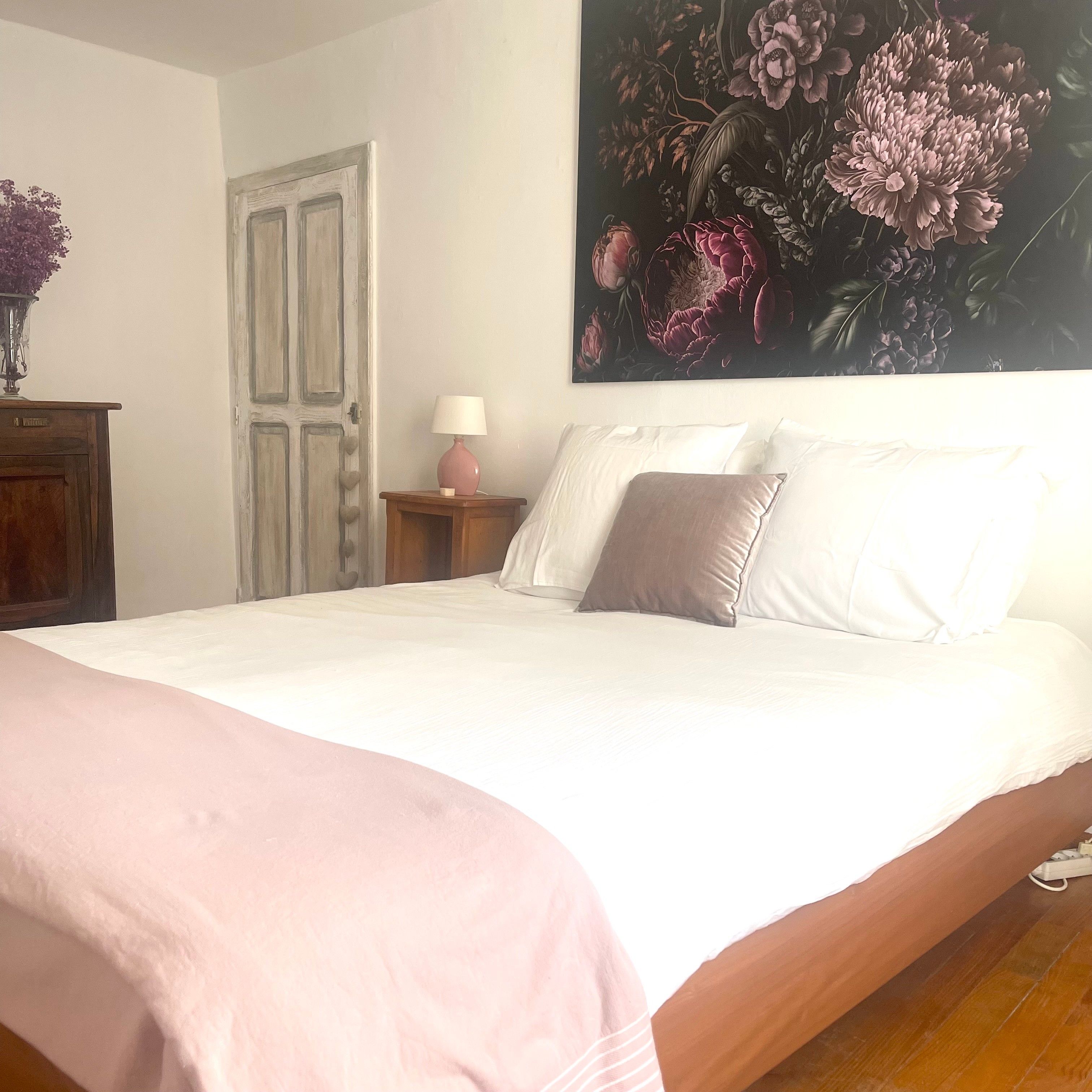 Un dormitorio con cama, una mesita de noche con lámpara, una puerta y una pared con un cuadro floral.