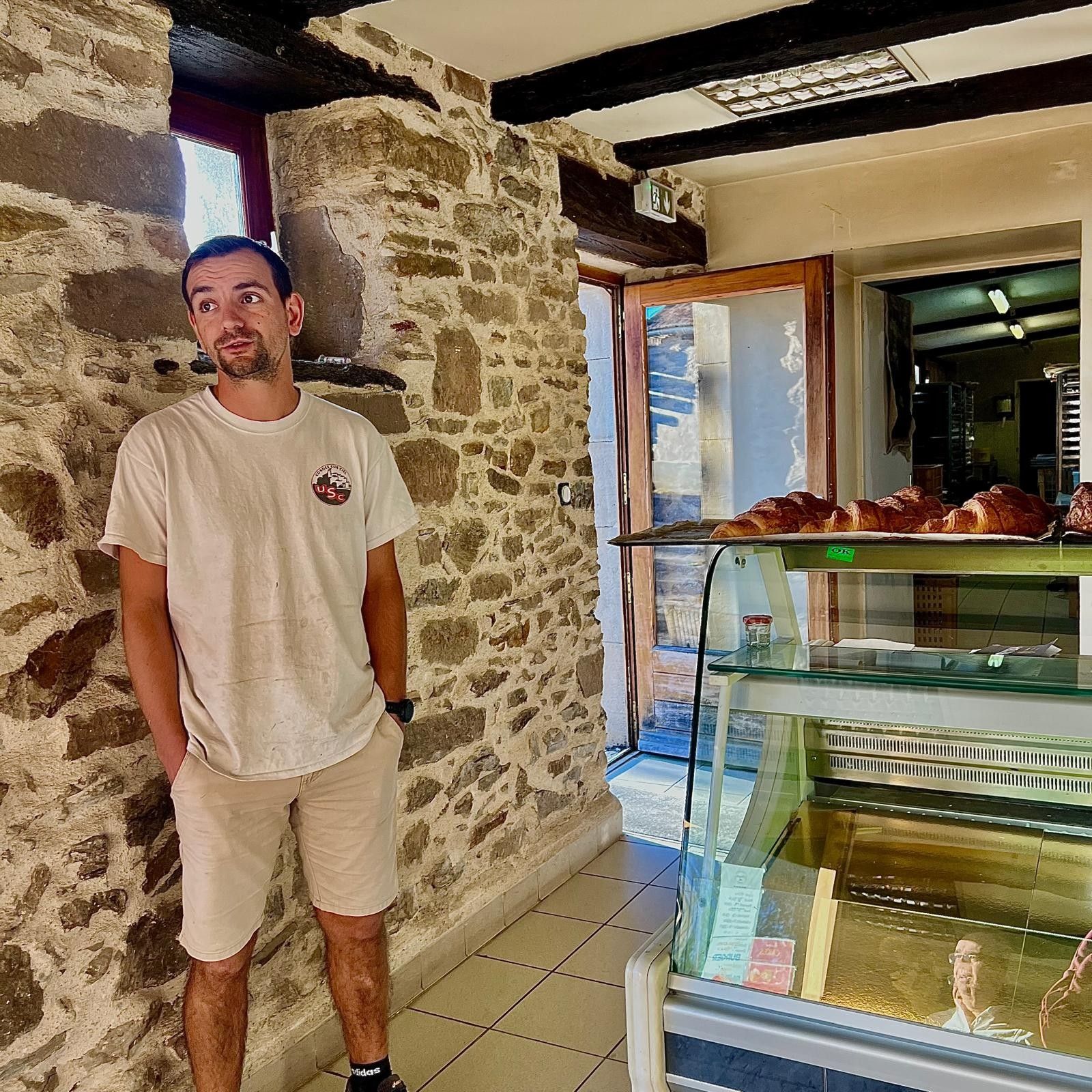 Un homme se tient dans un couloir aux murs de pierre, devant une vitrine de boulangerie remplie de pâtisseries.