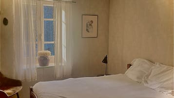 Une chambre à coucher comprenant un lit, une chaise, une fenêtre avec des rideaux, un tableau accroché au mur et un ventilateur de plafond.
