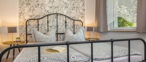 A bedroom with a bed, a nightstand, a window with curtains, and a view of greenery outside.