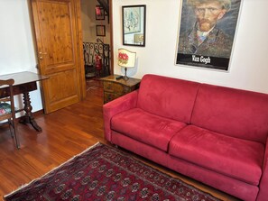 A room with a wooden door, a red sofa, a patterned rug, a small table with a lamp, and a framed picture on the wall. - House of the Hoopoe (Firenze)