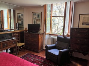 A room with a wooden dresser, a television on top, a chair, a radiator, and a window with a view of trees and buildings. - House of the Hoopoe (Firenze)