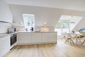 A modern kitchen with white cabinetry, a built-in oven, a microwave, and a coffee maker. There is a dining area with a table and chairs, and a large window providing natural light. - Holiday apartment with 1 bedroom (St. Peter-Ording)