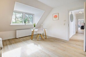 A room with a sloped ceiling, a desk with a chair, a radiator, a window with a view of trees, and a framed picture on the wall. - Holiday apartment with 1 bedroom (St. Peter-Ording)