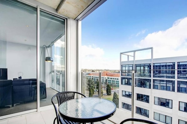 Parvekkeella on lasiovi, pyöreä pöytä ja tuoli, ja sieltä avautuu näkymä moderniin rakennukseen ja kirkkaalle taivaalle.
