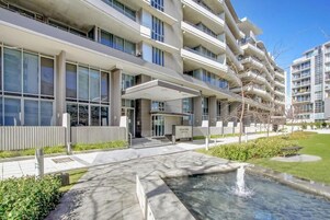 Un edificio moderno de varios pisos con un acceso cubierto, un jardín ajardinado y una fuente de agua.