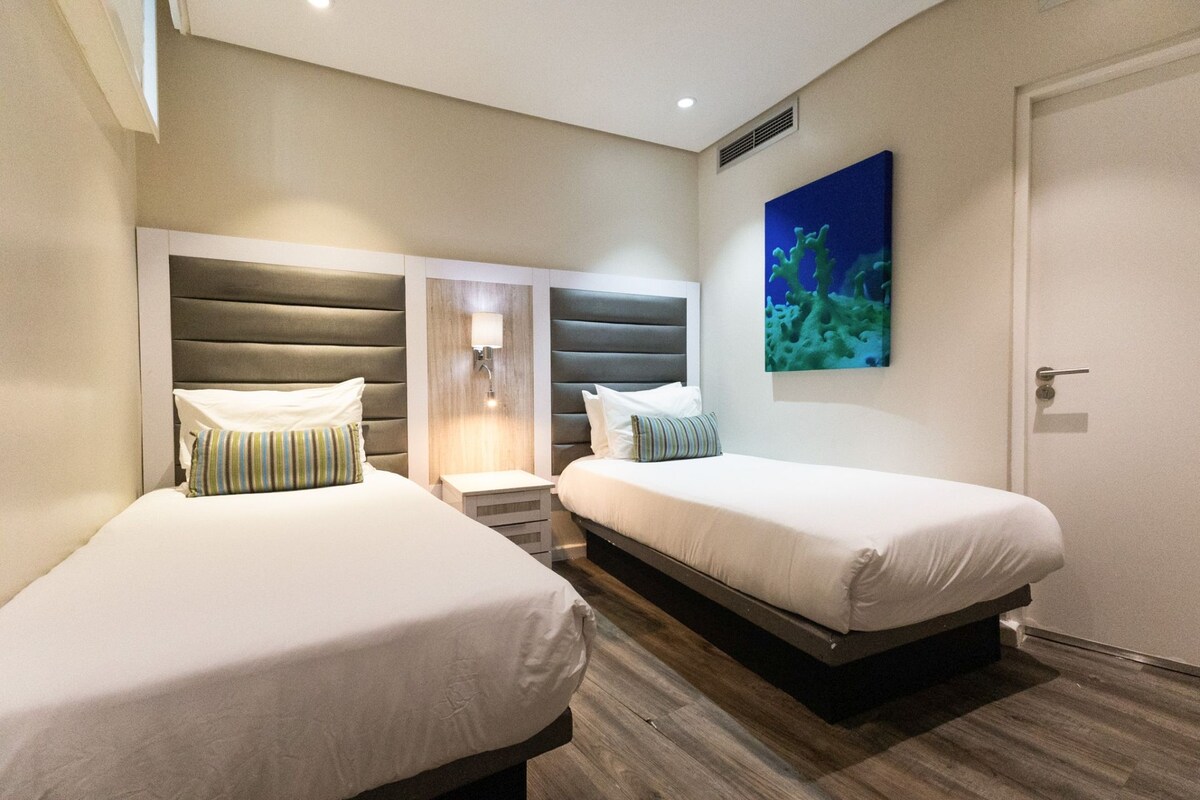 A hotel room with two beds, a nightstand, a wall-mounted light, and a painting of an underwater scene.
