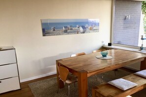 Ein Essbereich mit einem Holztisch, Stühlen und einer Bank. An der Wand hängt ein Bild und das Fenster bietet Blick auf den Strand.