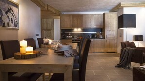 A modern kitchen with wooden cabinets, a dining table set for four, and a cozy seating area with a lamp. - Cgh Residences & Spas Le Lodge Hemera (Montvalezan)